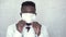 Front view a black male doctor in a white lab coat standing against a concrete wall, puts on and adjusts a medical mask