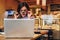 Front view. Business woman is sitting in cafe in front of laptop and looks at screen in surprise, lowering her glasses.