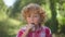 Front view close-up charming redhead little boy eating chocolate bar standing outdoors on sunny day in park. Portrait of
