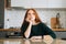 Front view of confused young woman writing handwritten letter sitting at table with envelope in kitchen room with modern