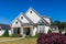 The front view of a cottage craftsman style white house with a triple pitched roof with a sidewalk, landscaping and curb