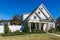 The front view of a cottage craftsman style white house with a triple pitched roof with a sidewalk, landscaping and curb