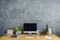 Front view of a desk with a computer, plants, notebooks and motorbike figure set on a cement wall. Product placement