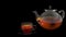 Front view of a glass teapot and a glass cup full of tea on glass dish on black background.