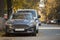 Front view of gray expensive luxurious car parked in quiet alley on sunny autumn day on blurred vehicles, walking people and