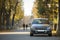 Front view of gray shiny empty car parked in quiet area on asphalt road at lamppost on blurred green and yellow trees