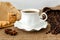 Front view of hot black coffee in white elegant cup with saucer, sweet cookies and scattering of coffee beans on rural sackcloth