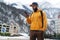 Front view. Man tourist in yellow hoodie, cap with backpack stands on background of high snowy mountains and using smartphone