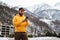 Front view. Man tourist in yellow hoodie, cap stands on background of high snowy mountains and using smartphone. Lifestyle