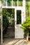 Front view of an old white glass greenhouse entrance with open wooden door. Inside garden isle with plants.