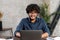 Front view of positive Hindi man in smart casual shirt using laptop while sitting at the desk in his flat. Young Indian