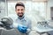 Front view of professional male dentist in white doctor coat and protective gloves sitting in dental chair and equipment, looking