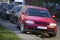 Front view of red car parked partly on sidewalk on background of long row of different vehicles along roadside on sunny autumn day