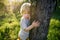 Front view of small boy standing outdoors in spring nature, hugging tree.