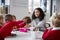 Front view of smiling female infant school teacher sitting at a table in the classroom with a group of schoolchildren