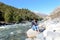 Front View of Solo Indian traveler in winter casual sitting alone near stream flowing water of remote mountain valley. Snow capped