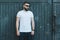 Front view. Young bearded hipster man dressed in white t-shirt and sunglasses is stands against dark wood wall.