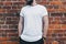 Front view. Young bearded millennial man dressed in white t-shirt is stands against dark brick wall. Mock up.