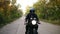 Front view of a young man in helmet with sunglasses and leather jacket riding motorcycle on a asphalt road in autumn