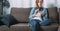 Front view of young woman in denim shirt sitting at home on couch and using smartphone. Girl uses digital gadget