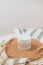 A frosted cosmetic glass jar for cream with a white lid stands on a cork stand on a white table, with dried flowers nearby. There