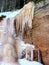 Frozen colorful icicles on the sandstone cliff wall, sunny day on the river bank.