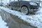 A frozen dirty car stands on the road in a city courtyard with puddles