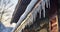 Frozen dripping water against ceiling lamp of house with stone wall. Spiked icicles at the edge of pitched gray roof with clumps