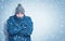 Frozen man in a blue jacket and hat stands against the wall, snow is falling around