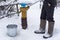 A frozen man in felt boots pours clean drinking water into a bucket using a water pump, against the backdrop of a snow-covered