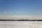 Frozen white snow lake on the background of the city with high-rise buildings and smoking huge pipes under a clear blue sky