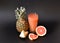 Fruit juice in a tall faceted glass on a black background, next to it are halves of ripe pineapple and grapefruit