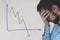 Frustrated business man standing near white board in office, covers his face with his hand at the falling graph of a stock market