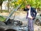 A frustrated guy stands near a broken car, looks under the hood and clings to his head. Outside.