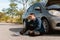 Frustrated man sitting near his broken car on the country road. Driver waiting for the insurance service. Broken car concept