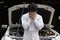 Frustrated stressed young mechanic man in white uniform covering his face with hands against car in open hood at the repair garage