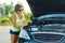 Frustrated woman driver stands near broken car on country road, looks at problem, an open hood