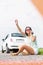 Frustrated woman hitchhiking while using cell phone on country road by broken down car