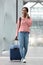 Full body happy young black woman walking with suitcase in airport terminal with cellphone