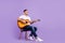 Full body photo of charming happy dreamy young man hold hands guitar sit char look empty space isolated on violet color