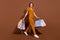 Full body photo of cheerful happy young woman hold bags walk empty space bargain isolated on brown color background