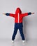 Full growth portrait of frolic blond kid boy in red and blue sport suit standing back to us with arms outstretched