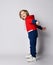 Full growth portrait of frolic blond kid boy in red and blue sport suit standing sideways to us with arms outstretched