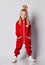 Full-growth portrait of stylish blonde kid girl in red jumpsuit and cool hairstyle standing with her hands in pockets