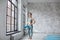 Full lengh attractive young woman with yoga mat, stands against window dresses stylish sportwear