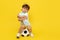 Full lengh photo of upset little boy sitting on soccer ball wearing headphone over yellow background.