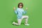 Full length body size view of attractive cheerful guy standing on knee showing horn sign isolated over bright green