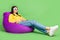 Full length body size view of pretty cheerful girl sitting in bag relax watching tv isolated over green color background