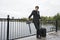 Full length of businessman with luggage leaning on railing along river