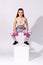 Full length fashionable sports woman in white tights and top holding pink dumbbells sitting in squat on cube and looking at camera
